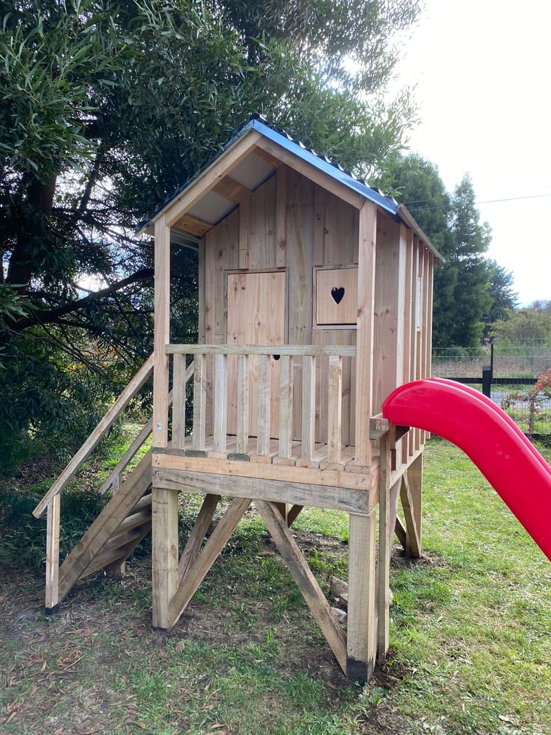 Casita de juegos de madera elevada, con escalera, baranda, ventana con corazón calado y tobogán rojo